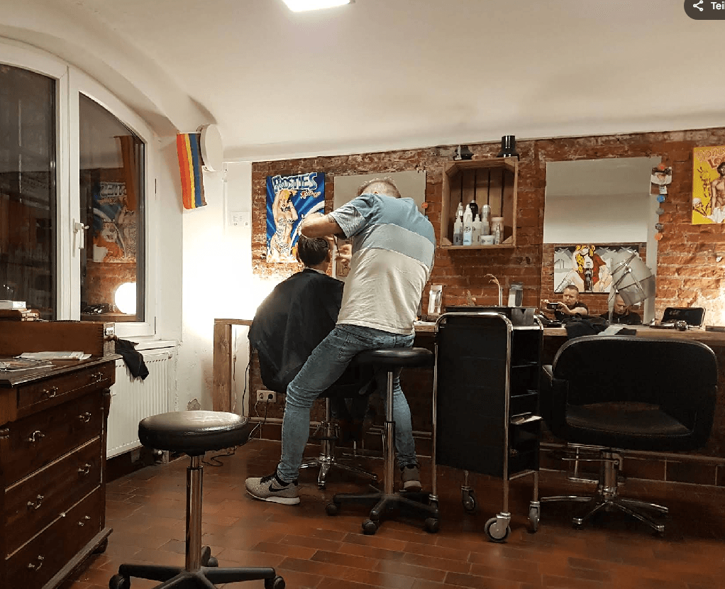 Friseur schneidet Haare in einem rustikalen Salon mit Backsteinwänden, Postern und einer Regenbogenflagge.